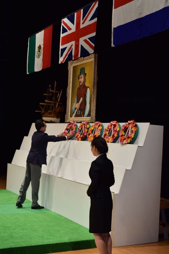 ４カ国の国旗と按針の肖像画が飾られたステージで花輪を奉呈する出席者代表＝市観光会館